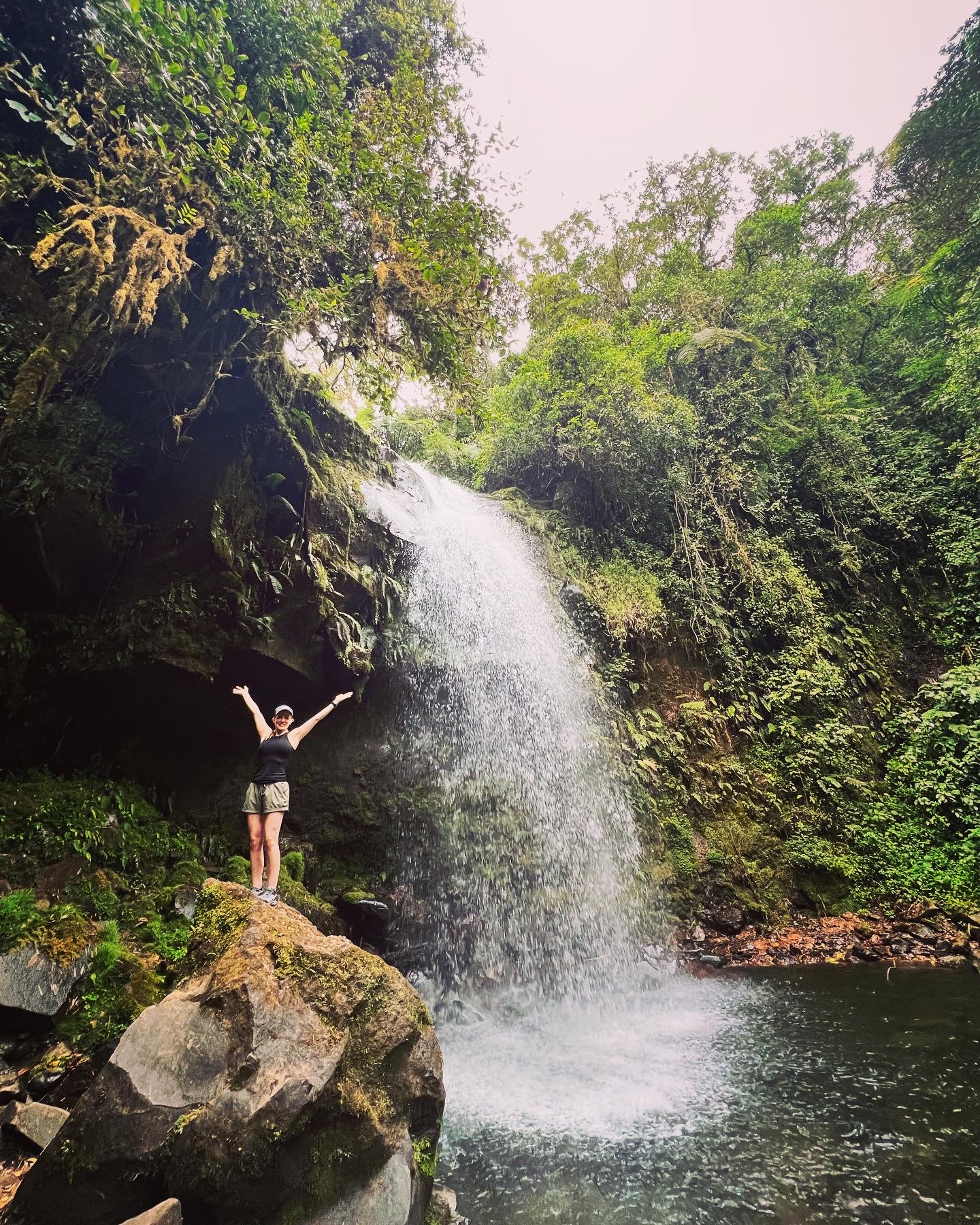 Tara Housh standing with arms outstretched on a mossy rock before a tall tropical waterfall in Boquete, Panama