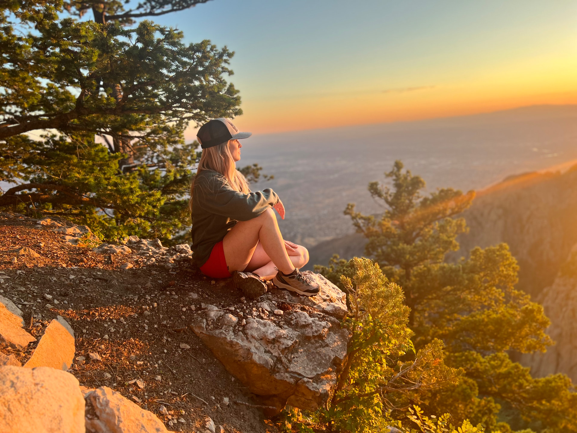 Woman seated on a mountain overlook at sunset, looking toward the horizon.